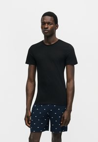 Homme portant un t-shirt noir cintré à manches courtes et un short bleu marine avec de petits logos blancs partout, debout devant un fond uni.
