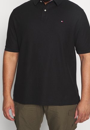 Hombre vistiendo una camiseta polo de manga corta negra con un pequeño logo rojo, blanco y azul en el pecho, combinado con pantalones cargo de color verde oliva.