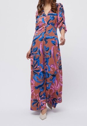 Femme portant une longue robe marron avec un imprimé floral abstrait bleu et rose, devant boutonné, manches trois-quarts froncées, et chaussures blanches pointues.