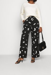 Pantalones anchos negros con un patrón de pinceladas blancas, combinados con un suéter de punto trenzado en crema texturizado y un bolso negro. Sandalias de tiras completan el look.