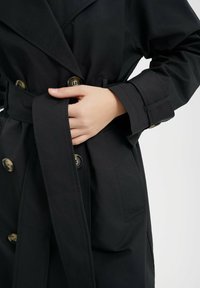 Czarny płaszcz typu trench, z podwójnym zapięciem, złotymi guzikami i materiałowym pasem. Tekstura wydaje się gładka z delikatnym połyskiem.