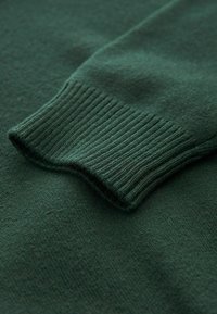 Close-up af en skovgrøn strikket sweaterærme med ribbet manchet på matchende stof baggrund.