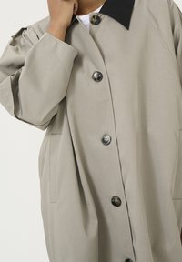 Beige oversized jas met een zwarte kraag, voorzien van grote knopen, zijzakken en een gladde textuur. Ontworpen met een comfortabele pasvorm.