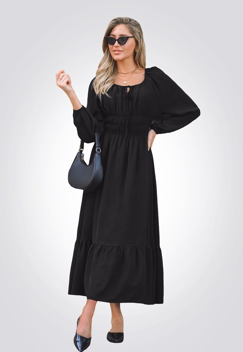 Vestido maxi negro con mangas largas, cintura fruncida y dobladillo con volantes. Combinado con un bolso negro y zapatos planos con punta. Presenta una textura sutil.