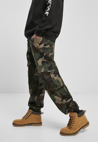 Camouflage cargo pants made of cotton blend, featuring multiple pockets, elastic cuffs, and a logo patch. Paired with tan boots.