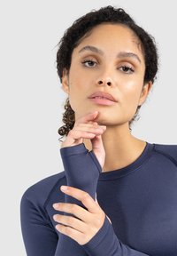 Marineblaues Langarm-Athletoberteil mit Daumenlöchern, glatter Textur und kontrastierenden Nähten. Das Modell posiert neutral, die Hände nahe dem Gesicht.