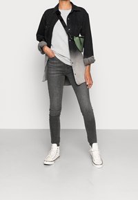 Giacchino in denim grigio e nero sfumato indossato sopra una camicia grigia chiaro con texture, abbinato a jeans aderenti grigio scuro e sneakers bianche alte.