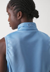 Svijetloplava bezrukavna košulja s glatkom teksturom, s preklopljenim kragnom i izvezenim "CK" logotipom na stražnjoj strani.