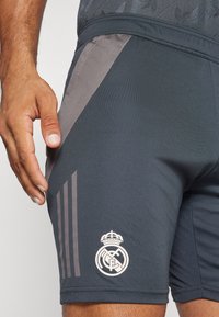 Pantaloni athletic grigi con una cintura testurizzata, pannelli laterali in rete grigia e logo del Real Madrid bianco. Presenta tre strisce verticali grigie.