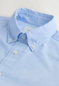 Chemise bleu clair à boutons blancs avec un col soigneusement plié sur un fond blanc.