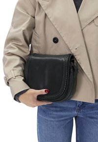 Zwarte leren schoudertas met een gestructureerde rand, ronde vorm en een ritssluiting, vastgehouden in een hand onder een beige trenchcoat.