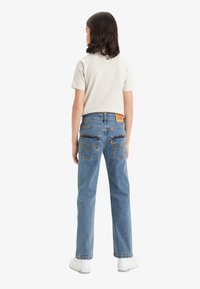 Jeans de mezclilla azul con un corte relajado, que cuentan con bolsillos traseros y un parche de cuero con la marca en la cintura. Combinados con una camiseta de color claro.