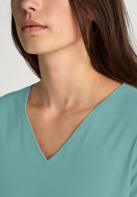 V-hals top van licht teal stof, met korte mouwen en een zachte textuur. Focus op de halslijn en de strakke ontwerpdetails.