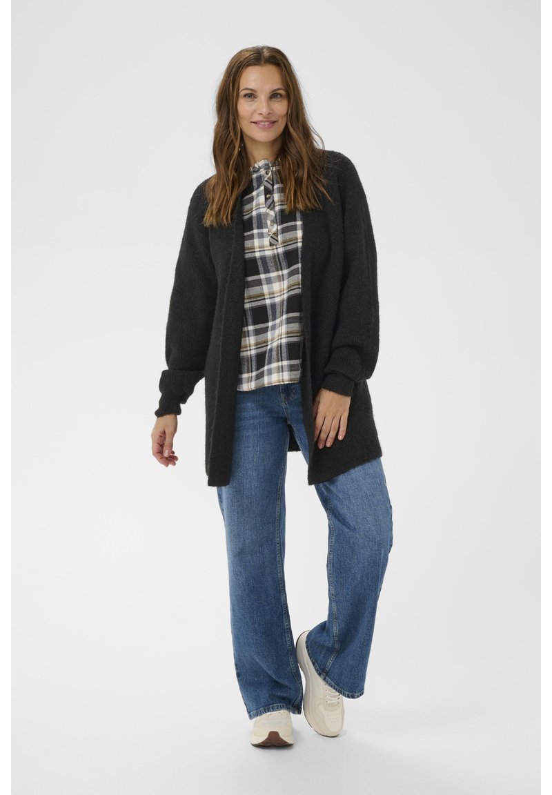 Zwart, gebreid vest met een losse pasvorm, gecombineerd met een geruit overhemd en blauwe jeans. Model draagt lichte sneakers en staat tegen een neutrale achtergrond.