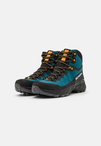 Bottes de randonnée avec un design noir et turquoise, présentant un dessus texturé, des semelles en caoutchouc, des lacets orange et un capot de protection renforcé.