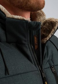 Veste vert foncé avec col en fausse fourrure, fermeture éclair et tissu texturé. Comprend un logo sur la poitrine et une couture détaillée.