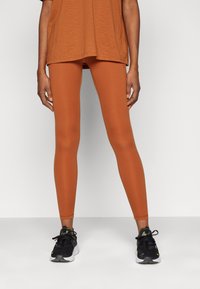 Leggings boje rđe napravljeni od glatkog i rastezljivog materijala, prilagođenog dizajna, s visokim strukom, u paru s crnim sportskim cipelama.