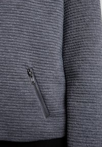 Suéter gris texturizado con patrón de canalé horizontal, que presenta un bolsillo con cremallera lateral con herrajes negros para un detalle funcional.