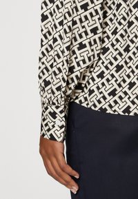 Long-sleeve blouse featuring a black and cream geometric pattern. Fabric has a slight sheen. Cuffed sleeve with a white button detail.