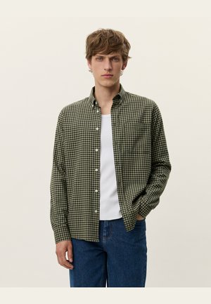 Chemise à carreaux verts à manches longues avec une poche poitrine et des boutons blancs, associée à un T-shirt blanc uni.