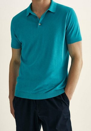 Man wearing a teal short-sleeve polo shirt and dark navy pants, standing with one hand in pocket against a plain background.