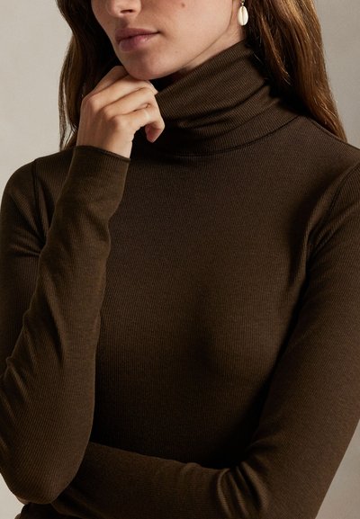 Mujer con suéter de cuello alto de canalé marrón oscuro y pendiente colgante con diseño de concha, mano descansando cerca del mentón sobre fondo neutro.