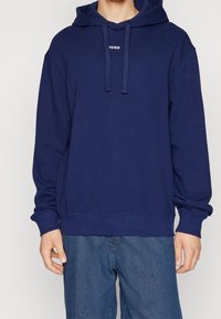 Námořnická modrá mikina s kapucí na stažení, žebrovanými manžetami a lemem. Na přední straně se nachází malé bílé logo "Hugo". Vyrobena z jemné, strukturované tkaniny.