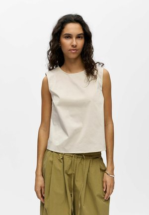 Femme aux cheveux bouclés et foncés, portant un haut beige sans manches et un pantalon ample vert olive, debout devant un fond blanc uni.