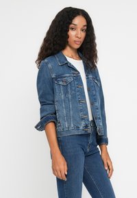 Chaqueta de mezclilla en azul medio con un diseño clásico, que cuenta con bolsillos en el pecho, cierres de botón y mangas remangadas para un ajuste relajado.