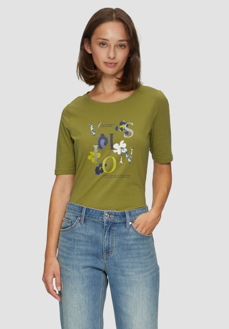 T-shirt olive vert à manches courtes avec un col rond, orné d'un imprimé graphique coloré de lettres et de fleurs. Porté avec un jean bleu.