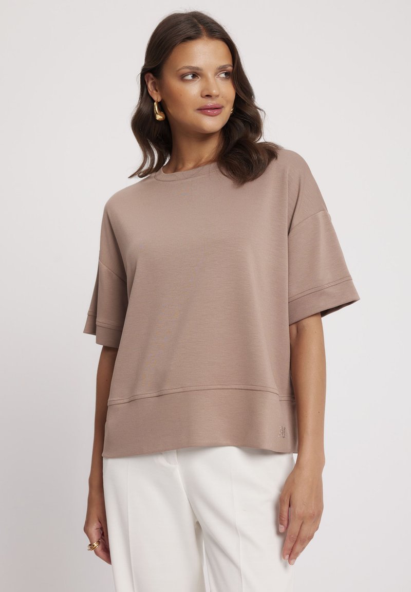 Jasnobrązowy, oversize, krótki top z okrągłym dekoltem, wykonany z fakturowanej tkaniny z delikatnym detalem na dolnej krawędzi. W połączeniu z białymi spodniami.