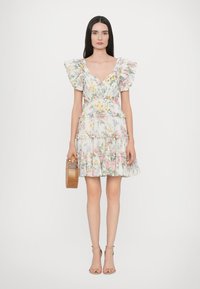 Abito con stampa floreale e maniche a balze, corsetto aderente e gonna a strati in colori pastello. Modella che tiene una borsa di legno e indossa scarpe con tacco.