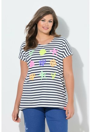 Uśmiechnięta kobieta o długich brązowych włosach nosi biało-granatową koszulę w paski z kolorowymi ilustracjami o tematyce morskiej oraz niebieskie spodnie.