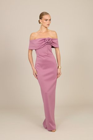 Femme en robe mauve longue jusqu'au sol, décolletée, avec un détail rose sur la poitrine, coiffée de boucles d'oreilles et d'un bracelet dorés, debout sur fond beige.
