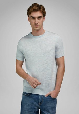 Jonge man met lichtbruin haar, gekleed in een lichtblauw gebreid shirt met korte mouwen en een blauwe spijkerbroek, poseert met één hand in zijn zak.