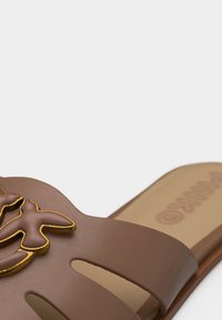 Gros plan sur une sandale à glissière marron ornée d'embellissements en forme d'oiseau bordés d'or sur une large bride et une semelle intérieure beige rembourrée.