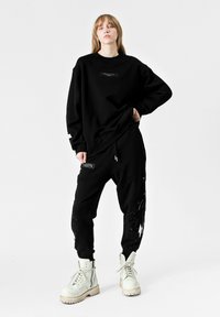 Sudadera negra con un detalle de bolsillo frontal, combinada con pantalones jogger negros que presentan un patrón texturizado, y botas de combate blancas.