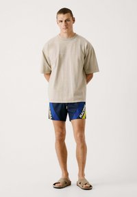 Bež majica oversize kroja s okruglim izrezom, u kombinaciji s mornarsko plavim kupaćim šortsem s žutim i bijelim dijagonalnim prugama i natpisom. Smeđe sandale.