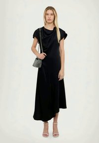 Vestido maxi preto com mangas curtas, design drapeado, textura suave e bainha assimétrica. Combinado com uma pequena mala xadrez.