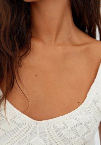 Haut en crochet blanc avec un décolleté en U, présentant des motifs complexes et une finition texturée, mis en valeur sur un teint de peau clair.
