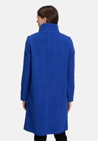 Kobieta z brązowymi włosami, ubrana w długi królewskoniebieski płaszcz z wysokim kołnierzem, stojąca tyłem na jednolitym jasnym tle.