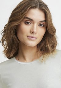 Světle šedé žebrované tričko s kulatým výstřihem; modelka má vlnité hnědé vlasy po ramena; malé kruhové náušnice; neutrální make-up.