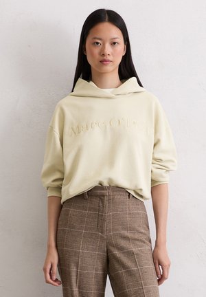 Sweat à capuche beige clair avec une coupe oversize, arborant un logo brodé. Associé à un pantalon sur mesure à carreaux marron avec des lignes et textures subtiles.