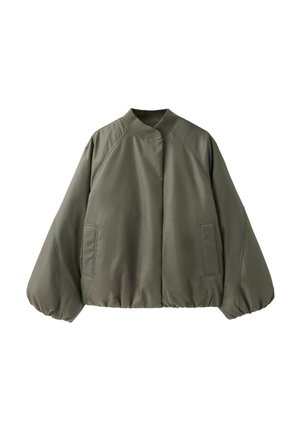 Ung kvinde med langt mørkt hår iført grøn bomberjakke, revnede blå jeans og beige vandresko, der sidder på en grøn metalbænk mod en murstensvæg.