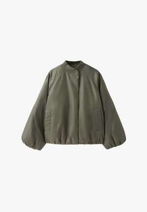Ung kvinde med langt mørkt hår iført grøn bomberjakke, revnede blå jeans og beige vandresko, der sidder på en grøn metalbænk mod en murstensvæg.