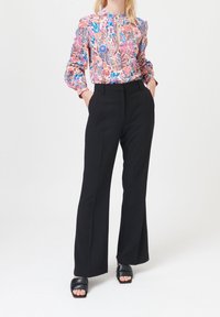Blouse fleurie en rose, bleu et vert sur un fond clair ; associée à un pantalon noir taille haute à jambes larges et des sandales noires à enfiler.