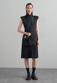 Costum negru personalizat cu o jachetă fără mâneci, cu umeri ascuțiți, pantaloni scurți cu cusături roșii contrastante și o geantă mică de umăr.