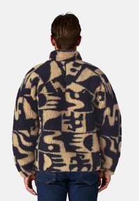 Giacca in fleece beige e blu navy con motivi astratti. Presenta un colletto alto e maniche lunghe, con una finitura testurizzata e soffice.