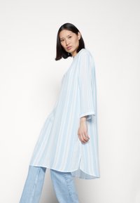 Túnica riscada em azul claro com mangas de três quartos, feita de um tecido leve, combinada com jeans de denim claro. Apresenta um ajuste descontraído.