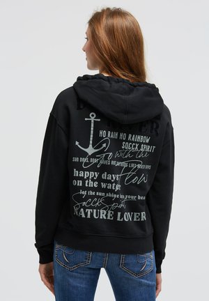 Woman with long hair wearing a black hoodie with anchor and nature-themed text, paired with blue jeans, facing away against a plain background.